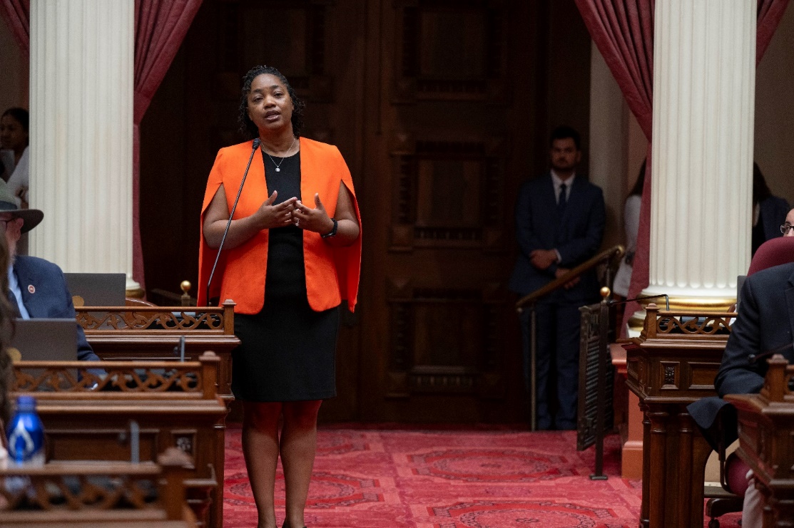 Wearing a red blazer and black dress, Senator Weber Pierson stands on the Senate floor to address the chamber. 